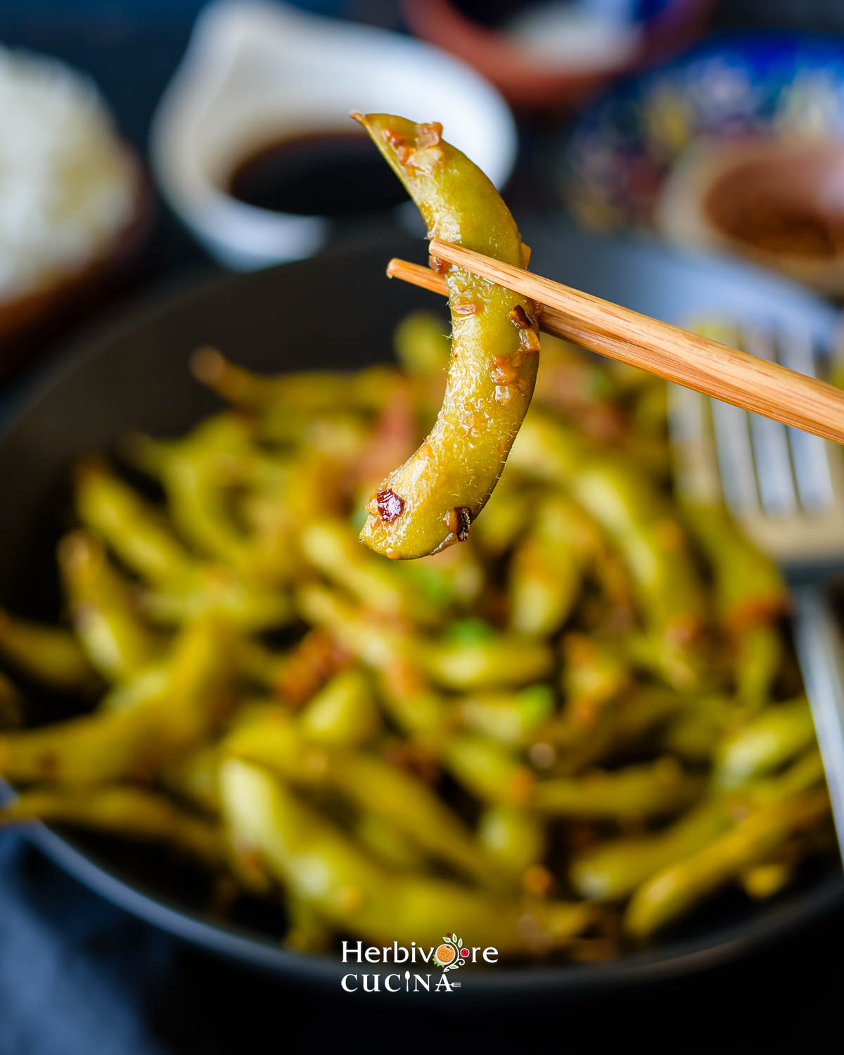 Holding an edamame pod in a chopstick over a bowl full of garlic edamame pods. 