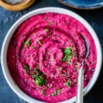 Roasted Beetroot Hummus in a white bowl with a spoon on the side and topped with sesame seeds, parsley and olive oil.