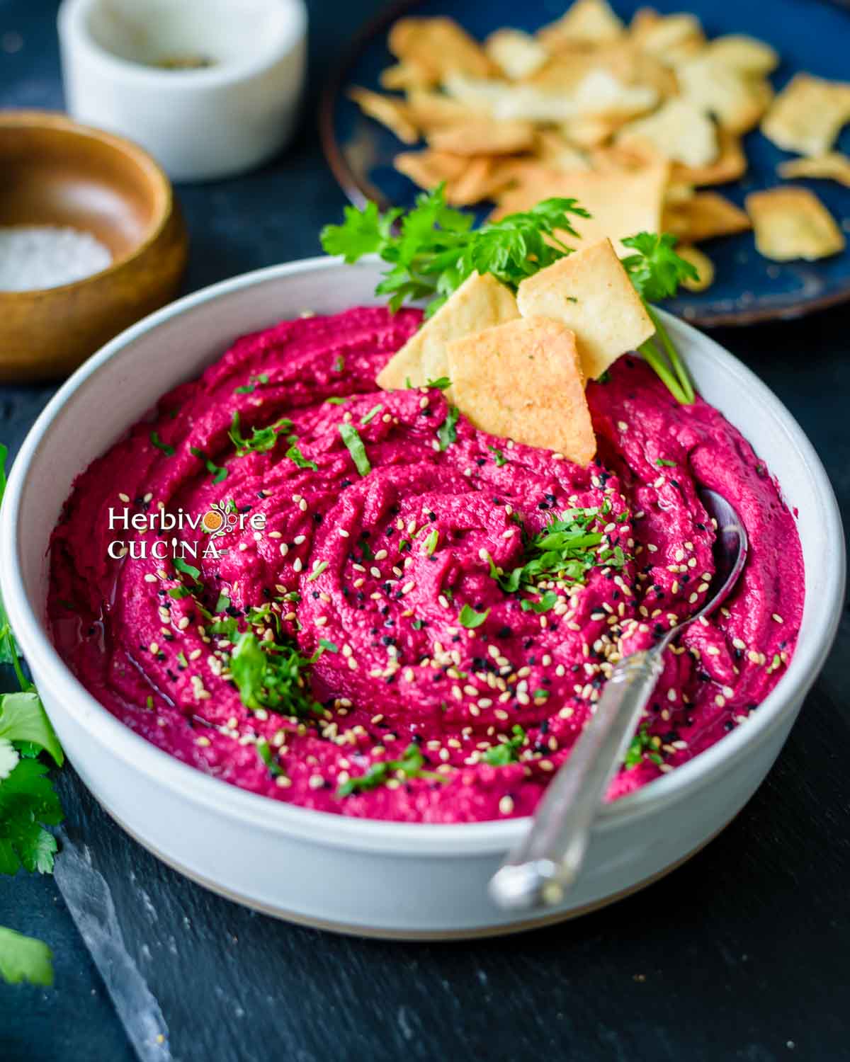 Roasted Beetroot Hummus in a white bowl topped with parsley, sesame and some chips. 