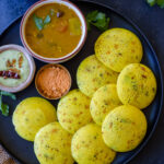 A black plate filled with Kanchipuram idli and served with chutneys and sambar.