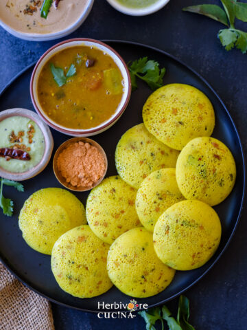 A black plate filled with Kanchipuram idli and served with chutneys and sambar.