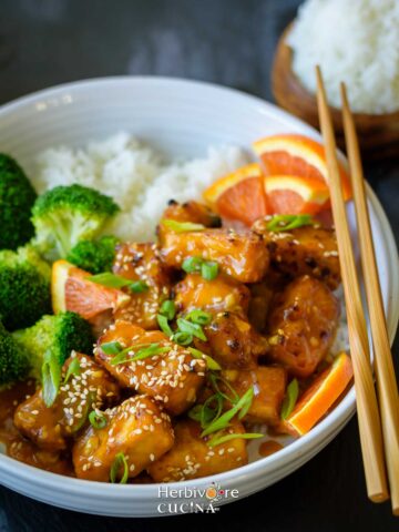 A bowl of orange tofu with steamed rice and broccoli on the side with a chopstick along with it.