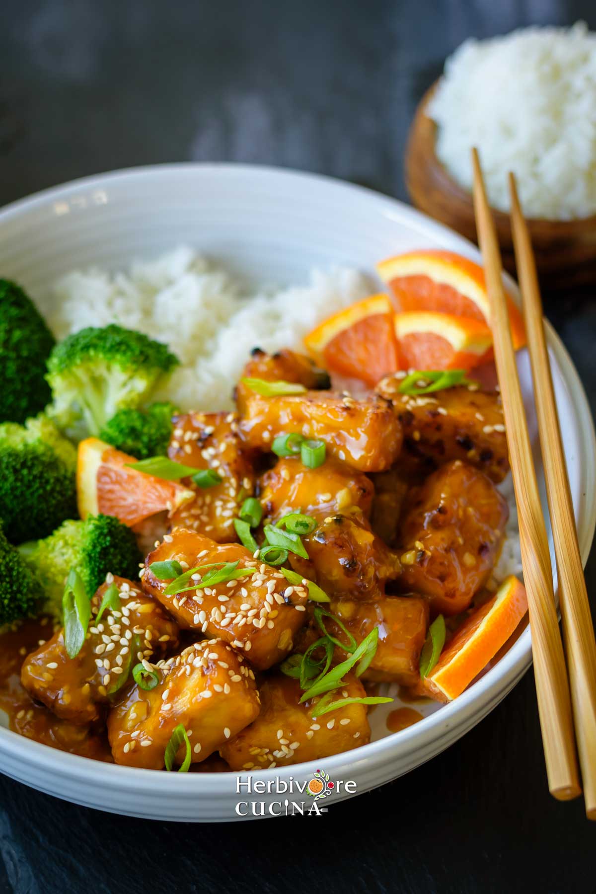 A bowl of orange tofu with steamed rice and broccoli on the side with a chopstick along with it. 