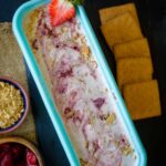 An ice cream tub filled with strawberry ice cream topped with a strawberry and graham crackers.
