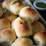 Baked vada pav in a tray topped with garlic chutney and green chili with chutneys on the side.