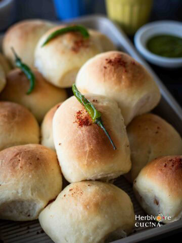 Baked vada pav in a tray topped with garlic chutney and green chili with chutneys on the side.