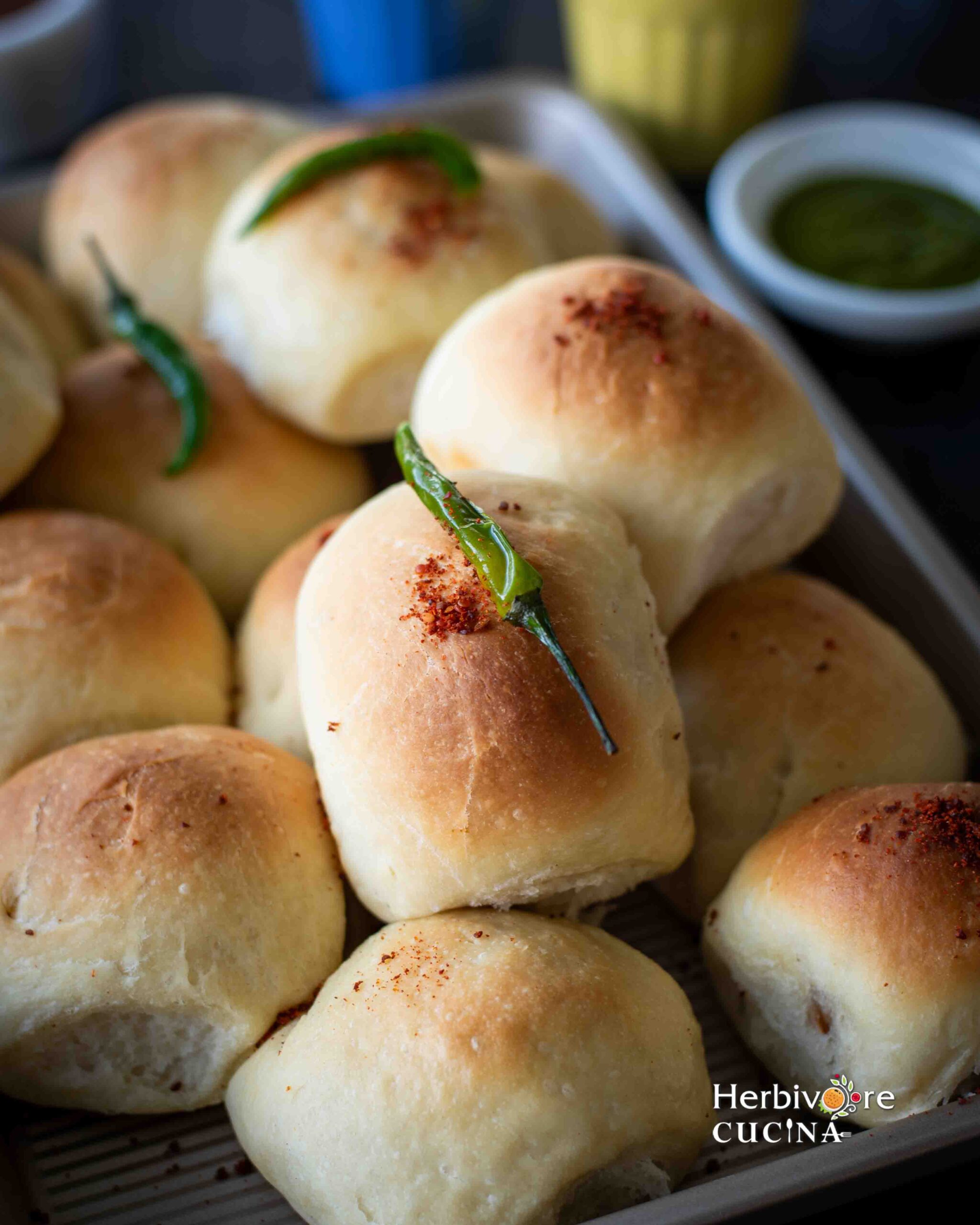 Baked vada pav in a tray topped with garlic chutney and green chili with chutneys on the side.
